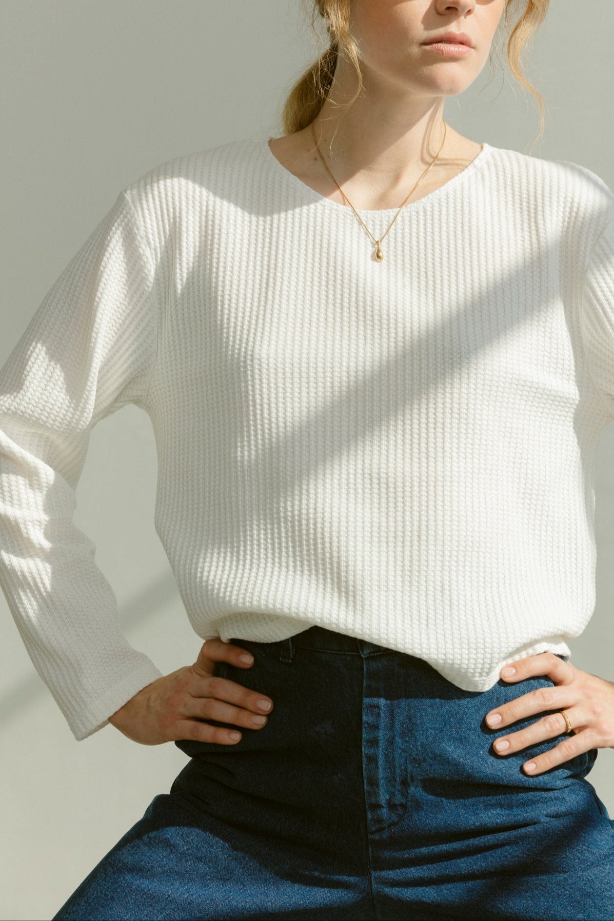 A woman wearing a white long-sleeve boatneck shirt and blue denim jeans, posing with her hands on her hips.